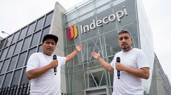 Dos hombres, uno con gorra, sostienen micrófonos y gesticulan frente a un edificio moderno de cristal con el logotipo de Indecopi en su fachada.