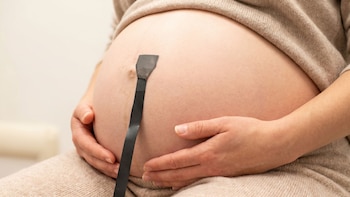 Primer plano del vientre de una mujer embarazada, con ambas manos apoyadas en la parte inferior y una tira negra, posiblemente un sensor, en la piel. Viste ropa beige.