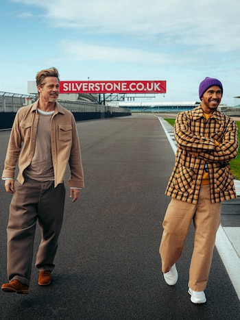 Brad Pitt y Lewis Hamilton caminan sobre el asfalto de una pista de carreras frente a un cartel rojo de Silverstone