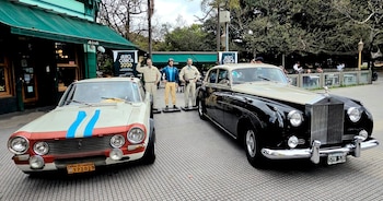 Dos autos antiguos, uno blanco con franjas azules y otro negro, junto a tres estatuas de pilotos y cartel de Autoclásica 2026