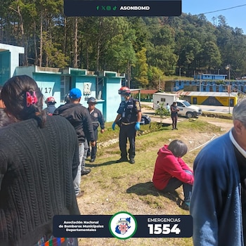 Bomberos y varias personas en un área verde con edificios azules y árboles al fondo. Una persona sentada en el suelo y otras de pie observando