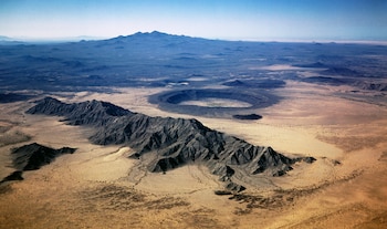 Vista aérea de un paisaje desértico con montañas rocosas, un gran cráter circular en el centro y terreno árido bajo un cielo azul claro