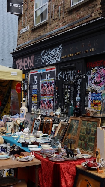 Un puesto de mercado callejero con mesas repletas de platos, vasos, jarras, figuras, cuadros y otros objetos, frente a un edificio con grafitis