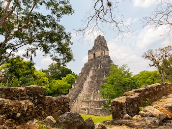 Pirámide de piedra con escalones rodeada de árboles frondosos y ruinas de piedra en primer plano bajo un cielo con nubes