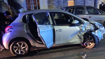 Automóvil plateado con frente y lado izquierdo severamente dañados, puertas abiertas y una persona junto al maletero en la calle de noche