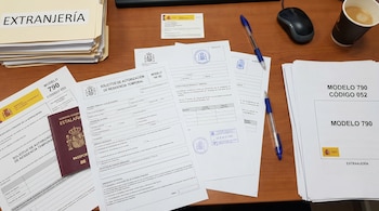 Mesa de madera con formularios de solicitud de residencia en España, un pasaporte, carpetas, bolígrafos, un ratón de ordenador y una taza de café.