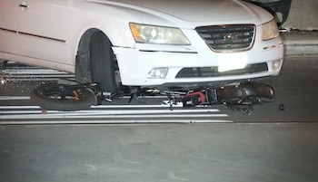 Un automóvil blanco de marca Hyundai está detenido con una bicicleta negra tirada debajo del parachoques delantero sobre el asfalto