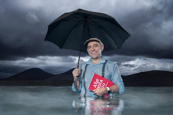 Hombre con boina y tirantes sumergido hasta la cintura en el agua, con un paraguas negro abierto en una mano y un libro rojo en la otra