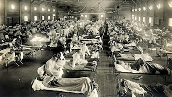 Gran sala de hospital con múltiples camas y pacientes. Enfermeras y personal médico asisten a los enfermos; algunos llevan mascarillas faciales