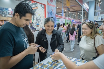 Tres personas adultas interactúan con tarjetas sobre una mesa blanca en una feria con stands y otras personas al fondo