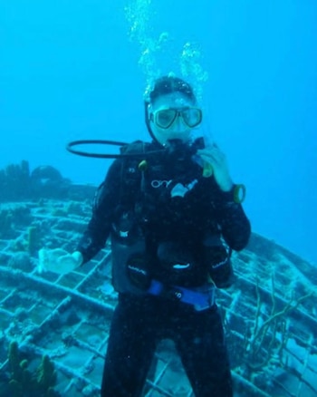 Rossana Almeyda, vestida con traje de buceo y máscara, emerge burbujas mientras bucea en el agua azul junto a una estructura submarina