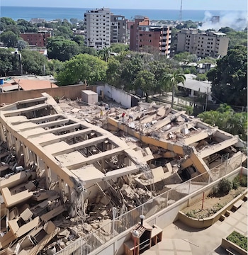 Edificio parcialmente derrumbado con escombros, tres personas sobre ellos, otros edificios y árboles al fondo, mar y cielo claro en el horizonte