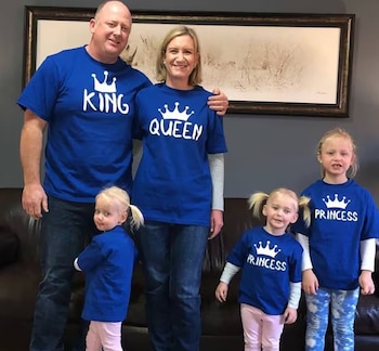 Hombre, mujer y tres niñas posan; visten camisetas azules con coronas y los textos King, Queen y Princess. Detrás hay un sofá y un cuadro
