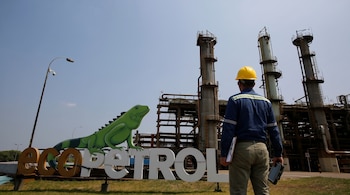 Un hombre de espaldas con casco amarillo y camisa azul mira el letrero de Ecopetrol con una iguana verde y torres industriales de fondo.