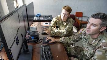 Dos hombres en uniforme militar están sentados frente a computadoras de escritorio, uno operando un ratón y el otro gesticulando mientras miran pantallas