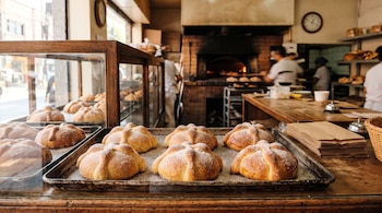 Panes con azúcar sobre una bandeja metálica, mostrador de madera con bolsas de papel y trabajadores junto a un horno al fondo.
