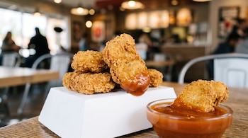 Nuggets de pollo frito apilados sobre una caja blanca junto a un tazón pequeño con salsa roja en una mesa.
