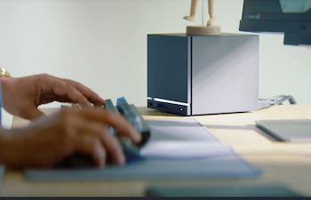 Manos de una persona escribiendo en un teclado, con una Steam Machine gris sobre un escritorio de madera en el fondo, junto a un monitor y una figura de madera
