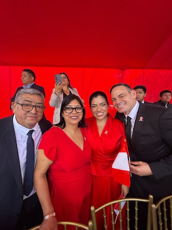 Cuatro personas sonríen. Dos hombres en traje, dos mujeres con vestidos rojos y pines de cinta. Un hombre sostiene bandera peruana. Fondo rojo liso