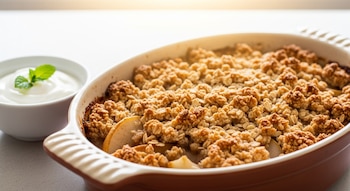 Crumble crocante de pera y manzana en un molde de cerámica ovalado, con un topping dorado y un tazón blanco con yogur natural y una hoja de menta al lado.