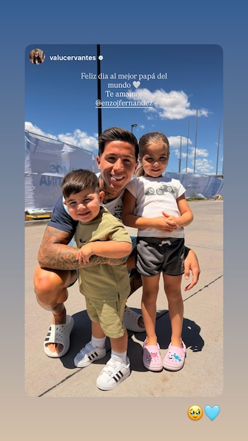Enzo Fernández agachado, sonriente, abrazando a un niño pequeño. Una niña sonríe de pie a su lado. Cielo azul con nubes y una valla al fondo