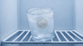 Un vaso de agua congelado con una moneda de 1 euro incrustada en el hielo, dentro de un congelador con las paredes y la rejilla escarchadas.