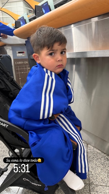 Un niño pequeño con chaqueta azul y franjas blancas sentado en un cochecito de bebé negro. Al fondo se observa un mostrador metálico de aeropuerto