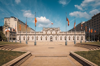 Vista frontal del Palacio de La Moneda, un edificio blanco neoclásico con una plaza pavimentada al frente y banderas ondeando bajo un cielo azul con nubes