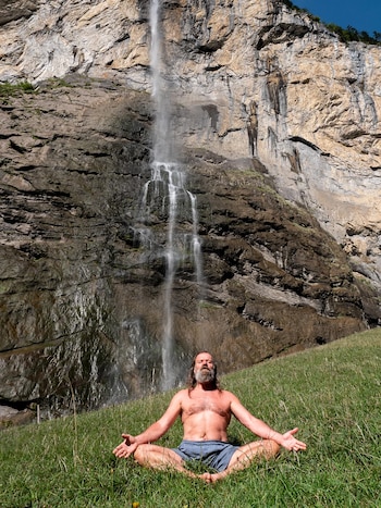 Hombre barbudo sin camisa meditando en hierba verde, con las piernas cruzadas y brazos extendidos, frente a una alta cascada que desciende por un afloramiento rocoso
