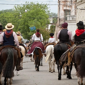 Varios jinetes con sombreros, boinas y vestimenta tradicional montan caballos de espaldas a la cámara por una calle