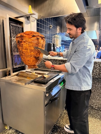 Luis Fonsi, de perfil, corta carne de un gran trompo de tacos al pastor con dos cuchillos en una parrilla de cocina. Viste una camisa de mezclilla azul