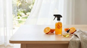 Botella rociadora con limpiador de naranja casero, naranjas, un cuenco con esencia y un paño sobre una mesa de madera frente a un ventanal con cortina.