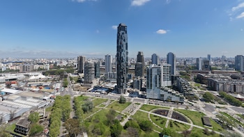 Vista aérea de un rascacielos oscuro, varios edificios residenciales, calles asfaltadas y un parque con senderos de diseño geométrico
