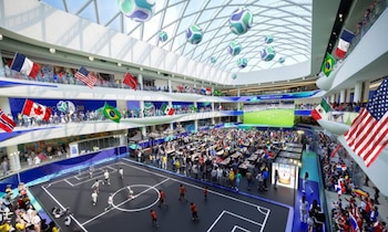 Vista aérea de un amplio interior con gente, una cancha de fútbol, banderas de países como EE.UU. y México, y pantallas gigantes mostrando un partido
