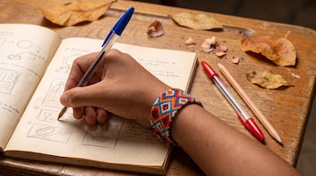 Primer plano de mano de estudiante escribiendo con bolígrafo azul en cuaderno abierto. Pulsera artesanal indígena colorida en la muñeca. Pupitre de madera con hojas y lápices.