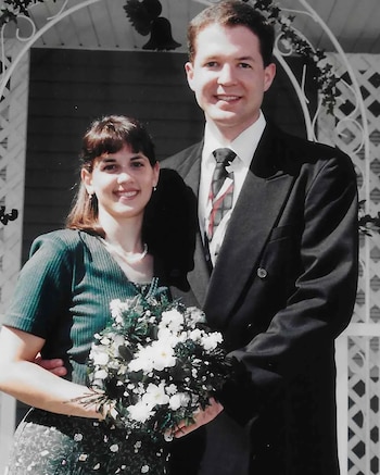 Un hombre y una mujer posan sonrientes a la cámara. Él lleva un traje oscuro, ella una blusa verde y un ramo de flores blancas. Detrás, un enrejado blanco