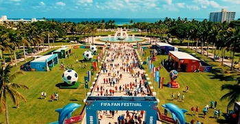 Vista aérea de un concurrido FIFA Fan Festival en Bayfront Park con personas, balones de fútbol gigantes, banderas y carpas en un parque verde con el océano al fondo