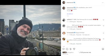 Primer plano de un hombre barbudo y sonriente con gorro negro y chaqueta oscura, sosteniendo una cámara. De fondo, un paisaje urbano borroso con edificios y un puente