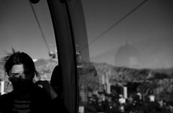 Fotografía en blanco y negro de una mujer en una cabina de teleférico con montañas y edificios visibles a través del cristal