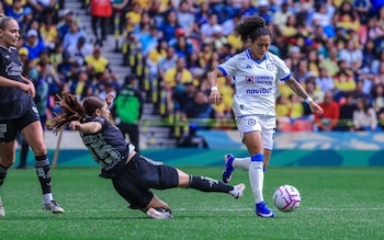 Una jugadora de Cruz Azul dribla el balón. Una jugadora de América Femenil se desliza en el césped. Una tercera jugadora. Público de fondo