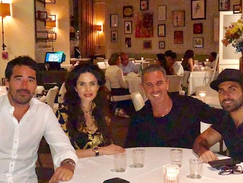 Cuatro personas, tres hombres y una mujer, sonríen sentadas a una mesa en un restaurante iluminado. Hay vasos vacíos y una vela encendida