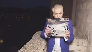 Hombre rubio con traje azul sostiene el álbum Pop Trash de Duran Duran que cubre su cara. Fondo oscuro con luces y muro de piedra