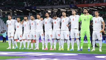 Once futbolistas iraníes, diez con uniformes blancos y uno (portero) de verde, de pie en un campo de fútbol con los brazos entrelazados, en un estadio