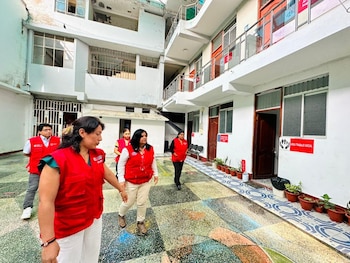 Varias personas con chalecos rojos del INABIF caminan por un patio interior de un edificio blanco con un piso de baldosas coloridas y macetas con plantas