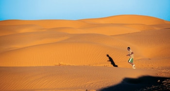 A visão de um homem correndo em uma duna de areia dourada sob um céu azul brilhante. Sua longa sombra é projetada na areia colorida