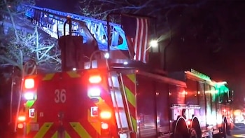 Parte trasera de un camión de bomberos rojo con el número 36 y luces de emergencia encendidas por la noche. Una escalera y una bandera estadounidense son visibles