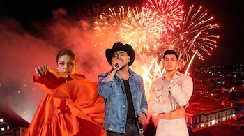 Una mujer, dos hombres (uno con sombrero y micrófono), fuegos artificiales rojos y dorados, y edificios de una ciudad en la noche.