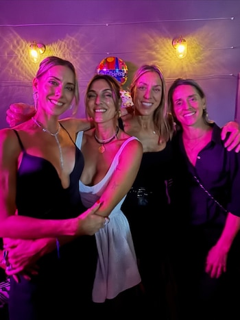 Cuatro mujeres sonrientes posan abrazadas bajo luces moradas y amarillas; un globo de cumpleaños y apliques de pared en el fondo