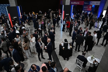 Vista aérea de una multitud de personas vestidas de negocios interactuando en un amplio salón de eventos con carteles del "AmCham Summit" y un área de bar