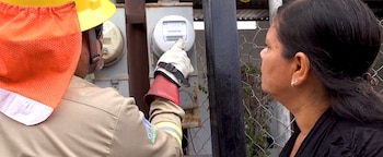 Un técnico con casco amarillo y guantes de seguridad blancos con rojo manipula un medidor de electricidad, mientras una mujer con pelo oscuro lo observa de perfil